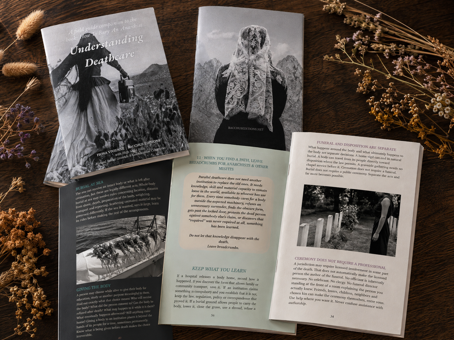 Several copies of Understanding Deathcare, a full-color Parallel Death Library field guide, arranged on a dark walnut table with dried wildflowers and grasses. The front and back covers appear alongside interior pages featuring black-and-white deathcare and burial photography, with sage, cream, and charcoal page designs.
